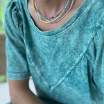 Woman wearing a teal shirt and three dainty bead necklaces. Necklaces are blues, white, gold and pinks.