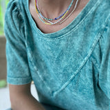 Woman wearing a teal shirt and three dainty bead necklaces. Necklaces are blues, white, gold and pinks.