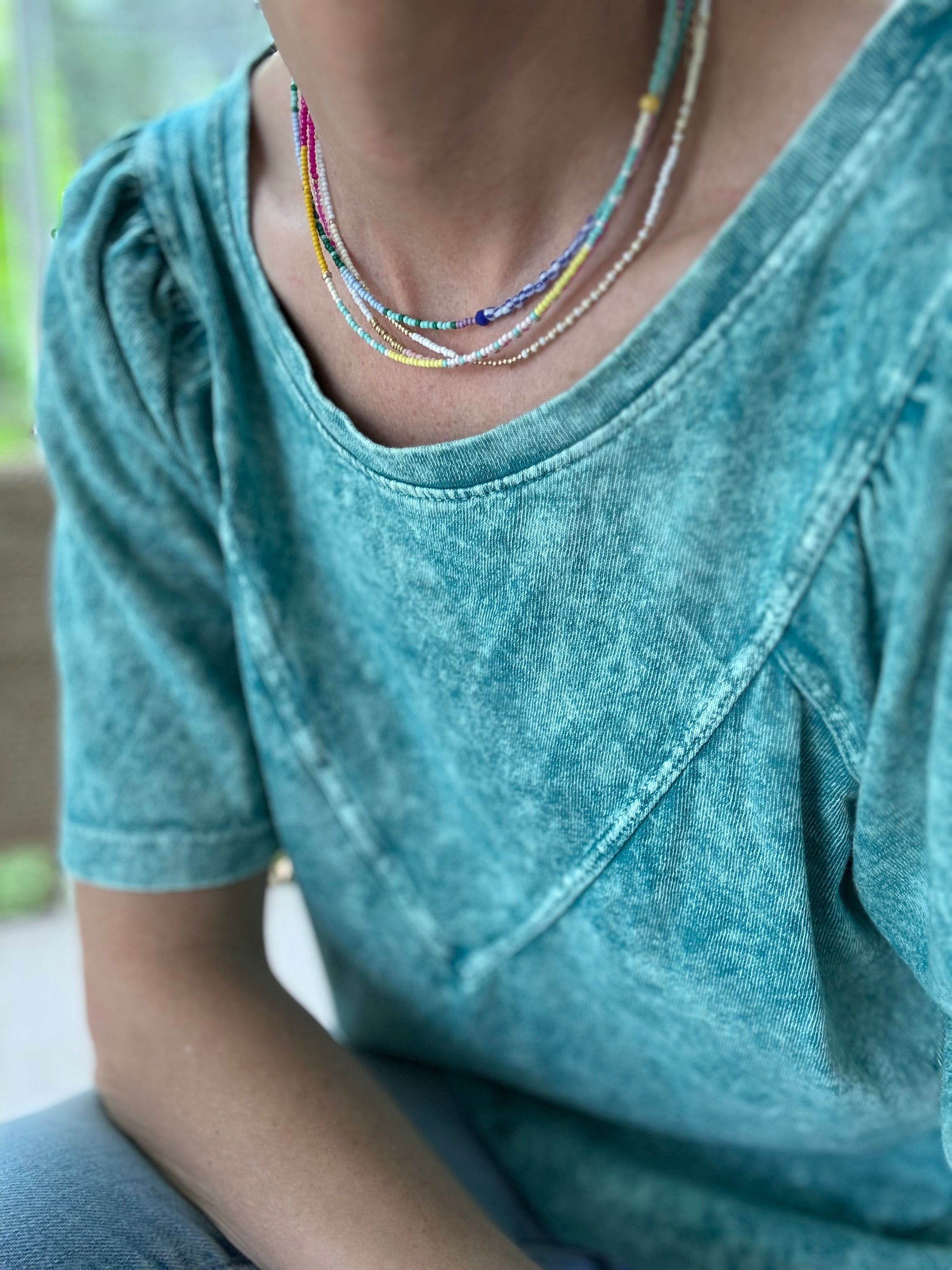 Woman wearing a teal shirt and three dainty bead necklaces. Necklaces are blues, white, gold and pinks.