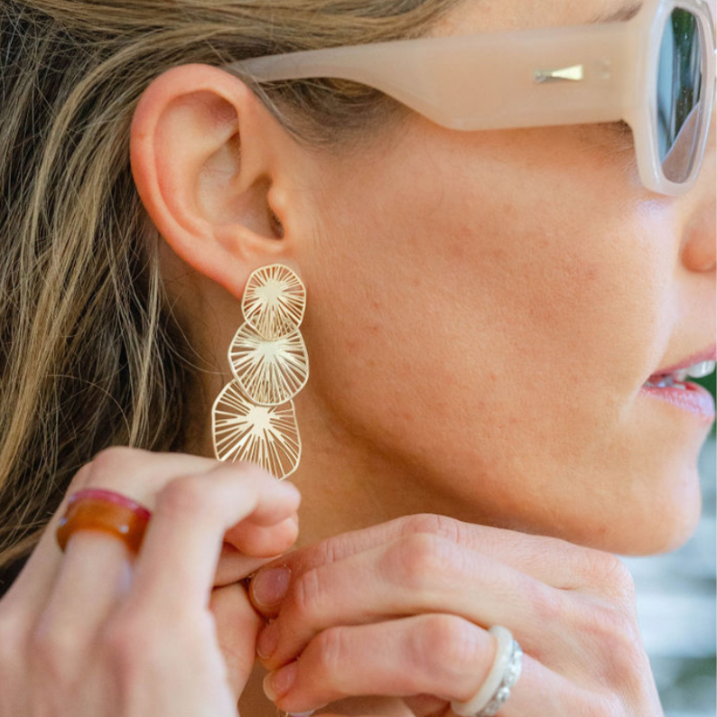 Woman wearing sunglasses reaching for her gold circular/stacked on each other sand dollar earring 