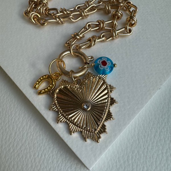 Picture of chunky gold chain necklace with a large gold heart pendant with a diamond stud in the center. Necklace also has two other charms attached, one a golden horseshoe and the other a blue bead with a flower design.