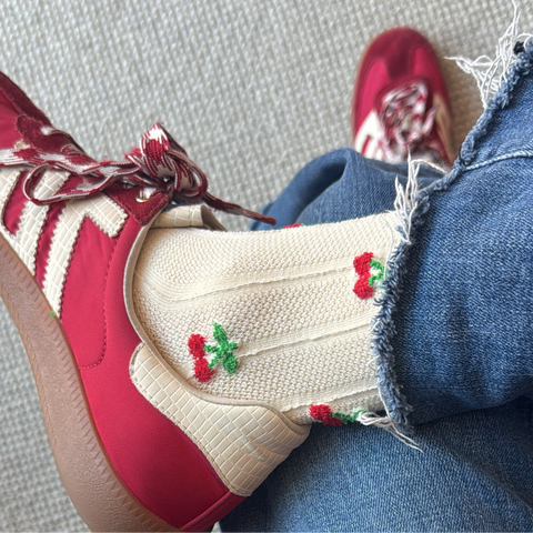 Picture of crossed legs wearing jeans and red sneakers. Socks are cream colored with small cherries on them.