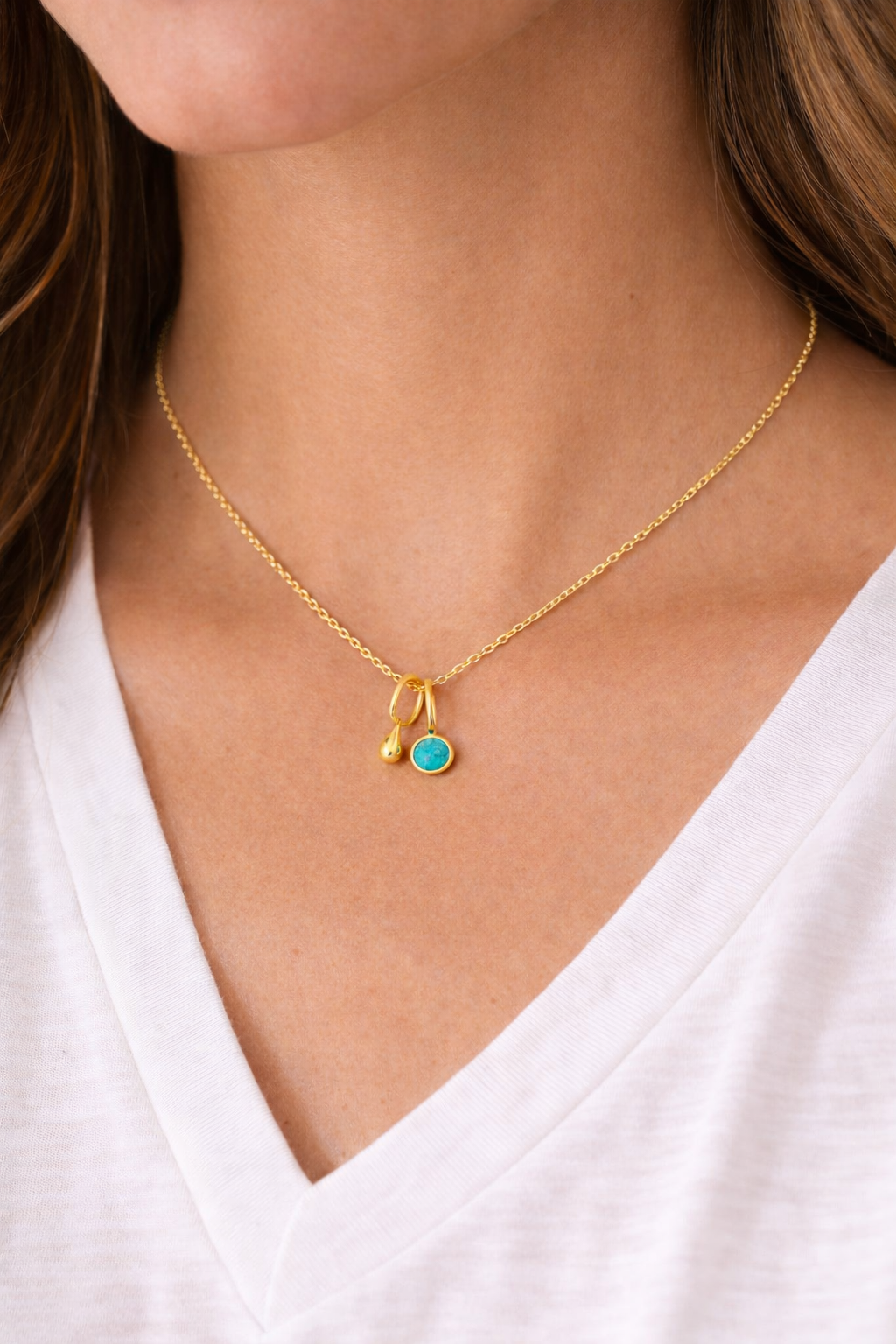 Gold necklace with a turquoise pendant and a small gold teardrop pendant on a person wearing a white top.