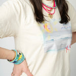Picture of a woman wearing a cream colored t shirt with a picture of a beach scene. She also is wearing a layered stack of beaded necklaces in bright coral, light blue and a gold toggle heart necklace. She is also wearing a beaded turquoise and yellow bracelet bundle.