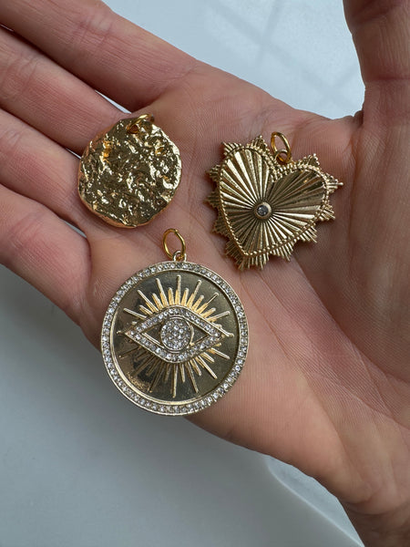 A person holding three different gold medallions: a circular melted gold, a gold heart shape, and a gold circle with an eye in the middle with shiny diamonds around it 