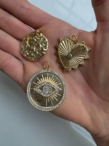 A person holding three different gold medallions: a circular melted gold, a gold heart shape, and a gold circle with an eye in the middle with shiny diamonds around it 