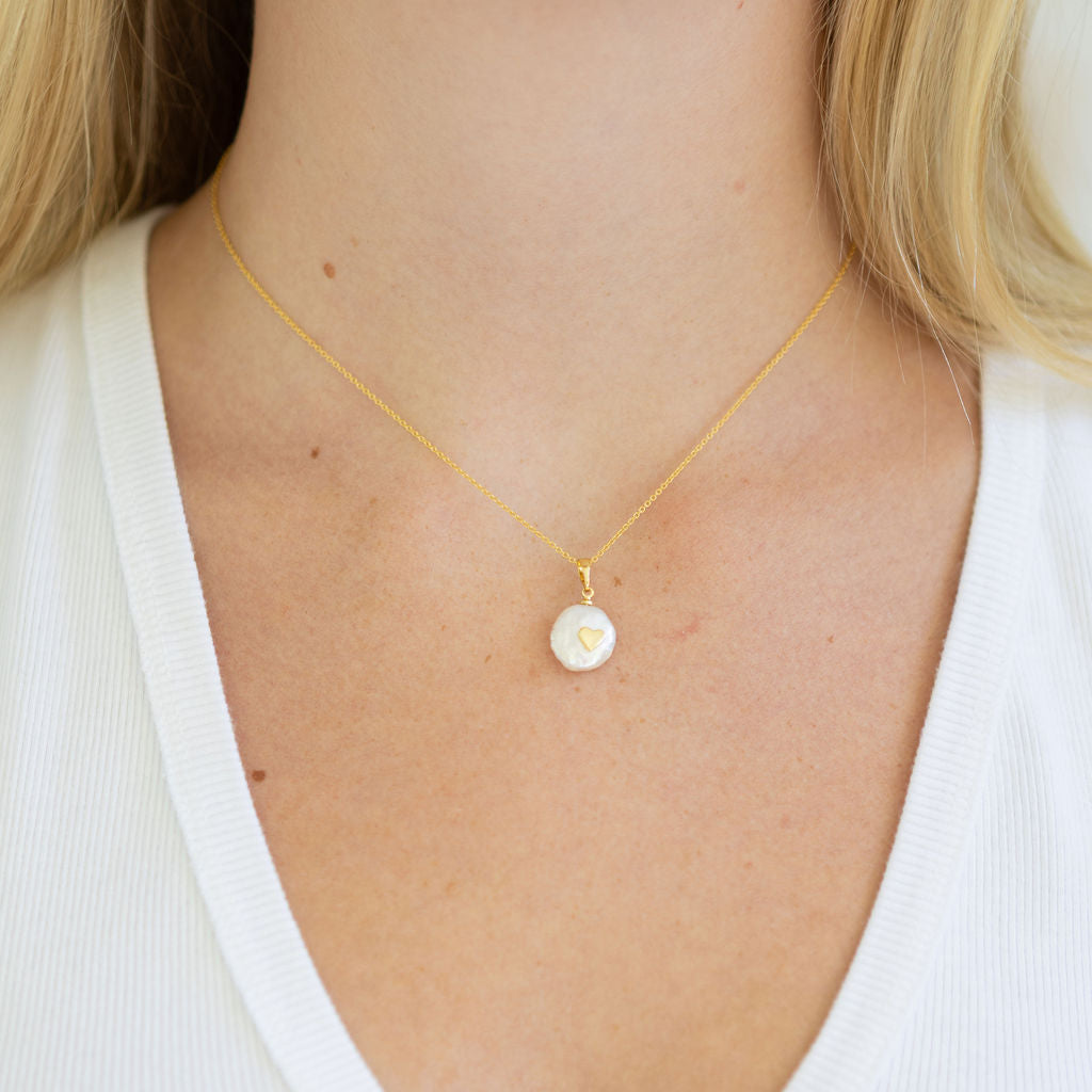 Delicate gold chain with a small pearl disc with a tiny gold heart on it worn by a person in a white shirt.