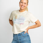 Happy blonde woman wearing a cream colored t shirt. T shirt has a beach scene printed on the front of it with the letters XOXO in pink.