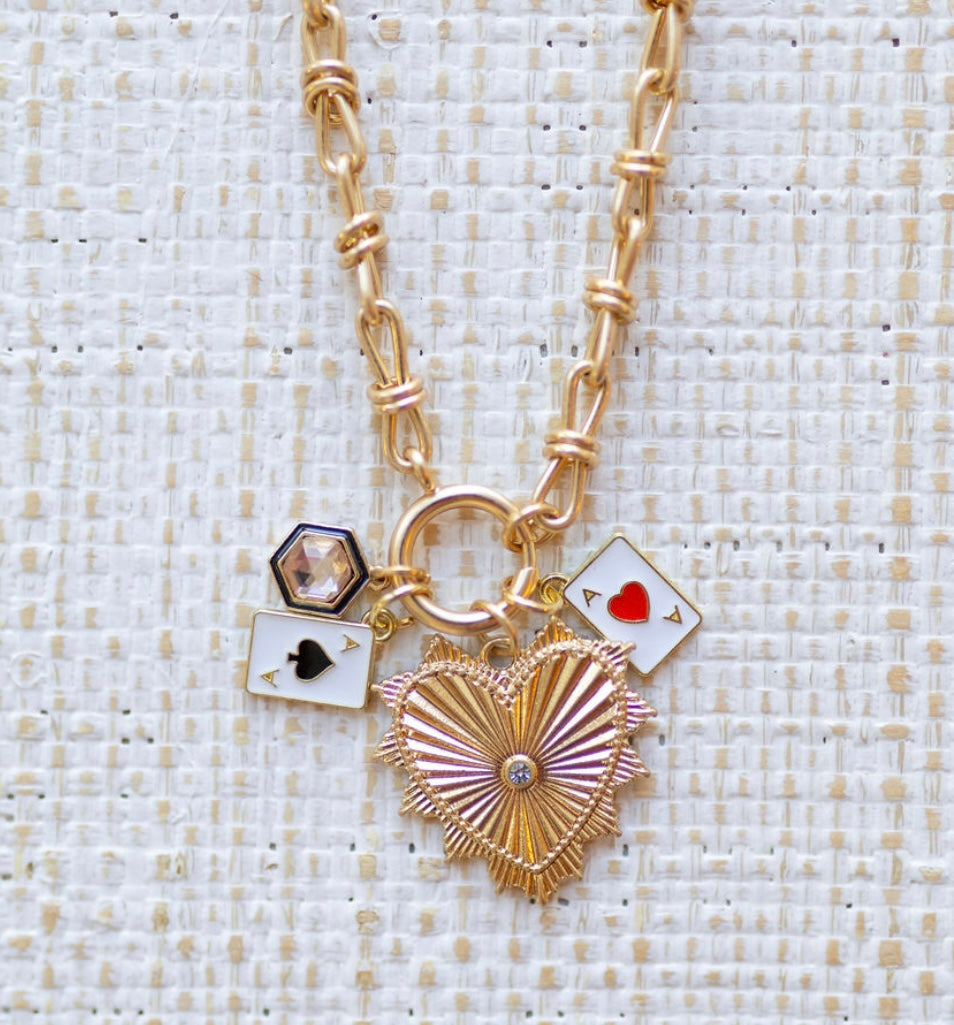 Picture of gold toggle chain with a large gold heart pendant with a diamond in its center,a blue bordered diamond charm, a an ace of spades charm and an ace of hearts charm.