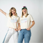 Picture of two women wearing jeans and matching beige cropped t shirts that have a beach scene. Women appear to be very happy and are accessorizing with trucker hats, sunglasses, statement jeans and beaded necklaces.