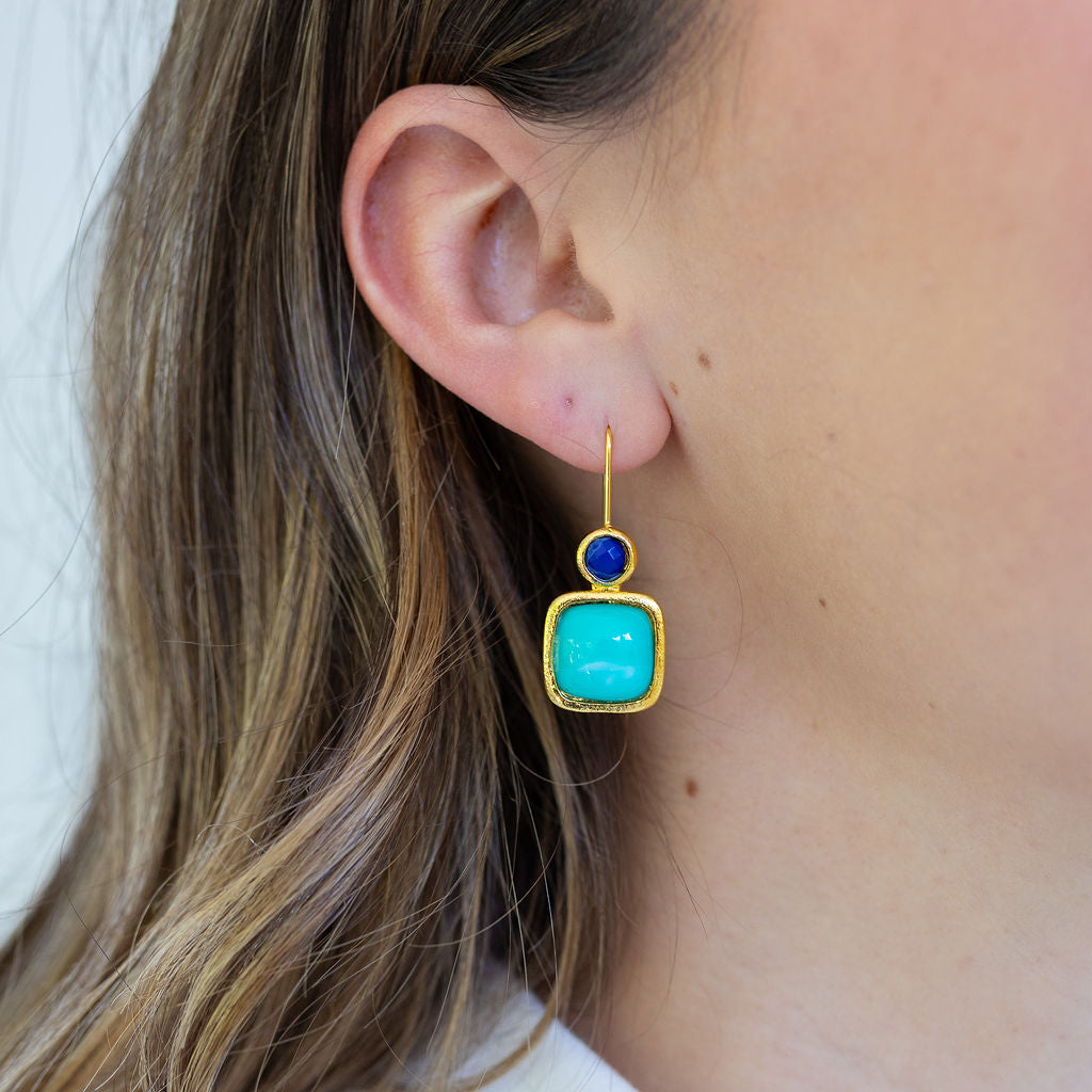 Picture of a woman wearing dangling earrings with light blue squares topped by tiny dark blue beads.