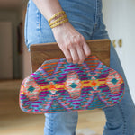 Person holding in their hand a clutch with brown at the top and a purple, orange, and yellow colored cloth design 