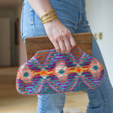 Person holding in their hand a clutch with brown at the top and a purple, orange, and yellow colored cloth design 