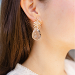 Woman wearing a large dangle earrings with a sunburst gold stud and large faceted teardrop crystal beneath.