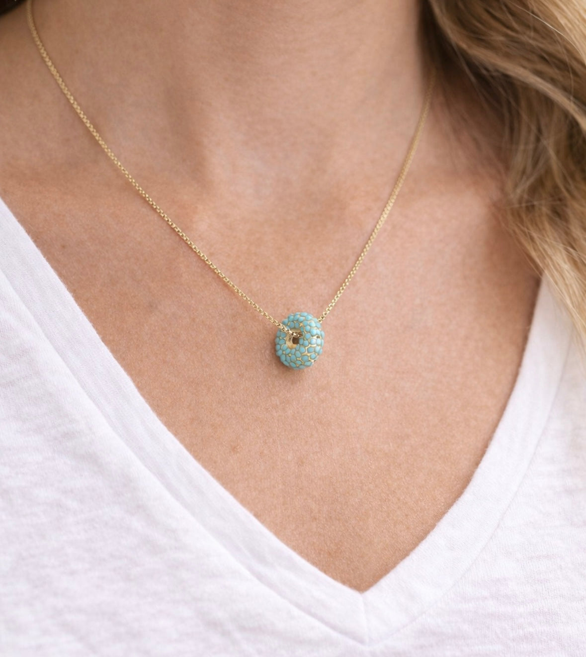 Gold necklace with a donut shaped turquoise pendant worn by a person in a white shirt.