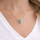 Gold necklace with a donut shaped turquoise pendant worn by a person in a white shirt.