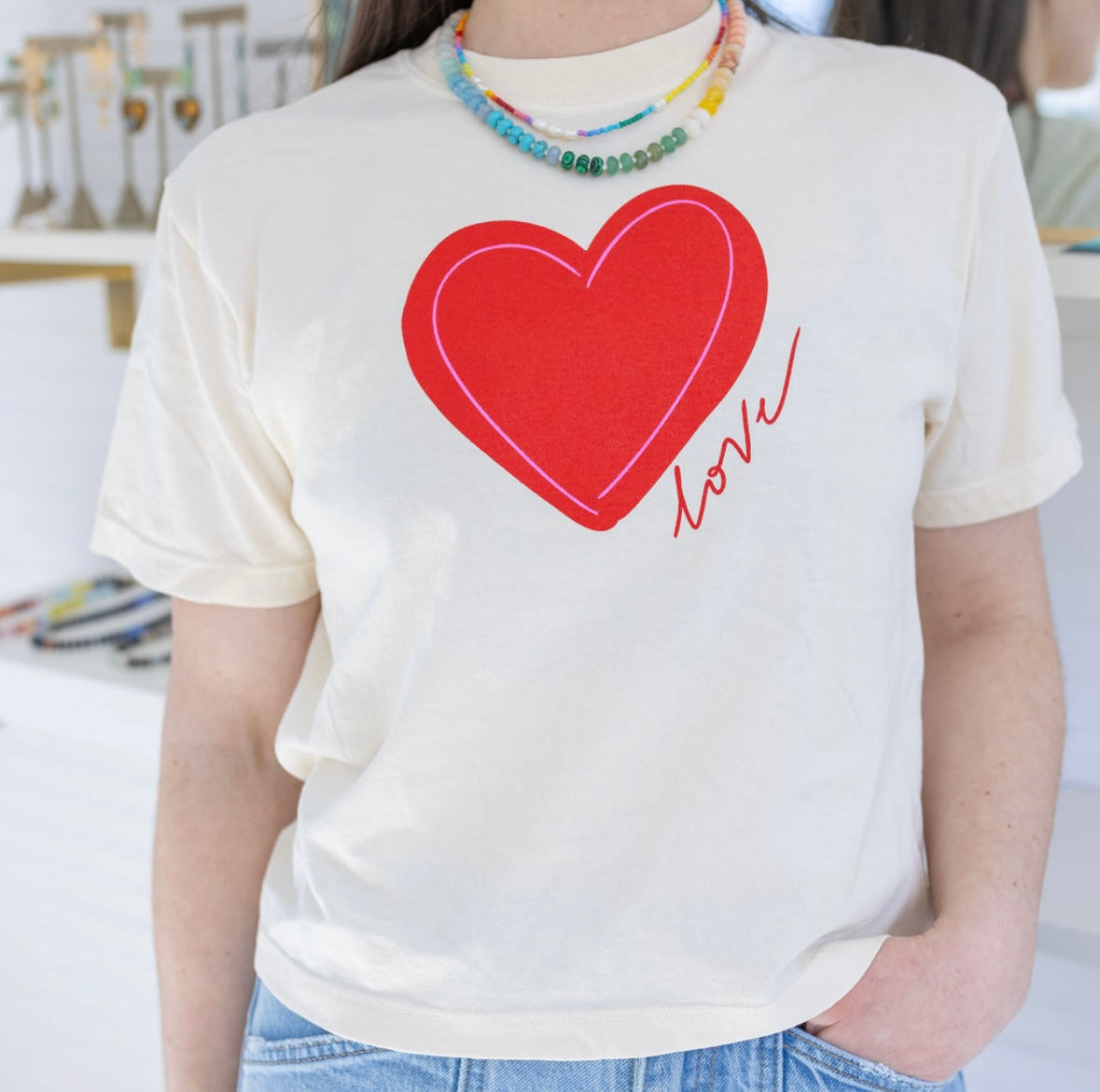 Woman wearing a a beige Tshirt with a red heart and script that reads "love".