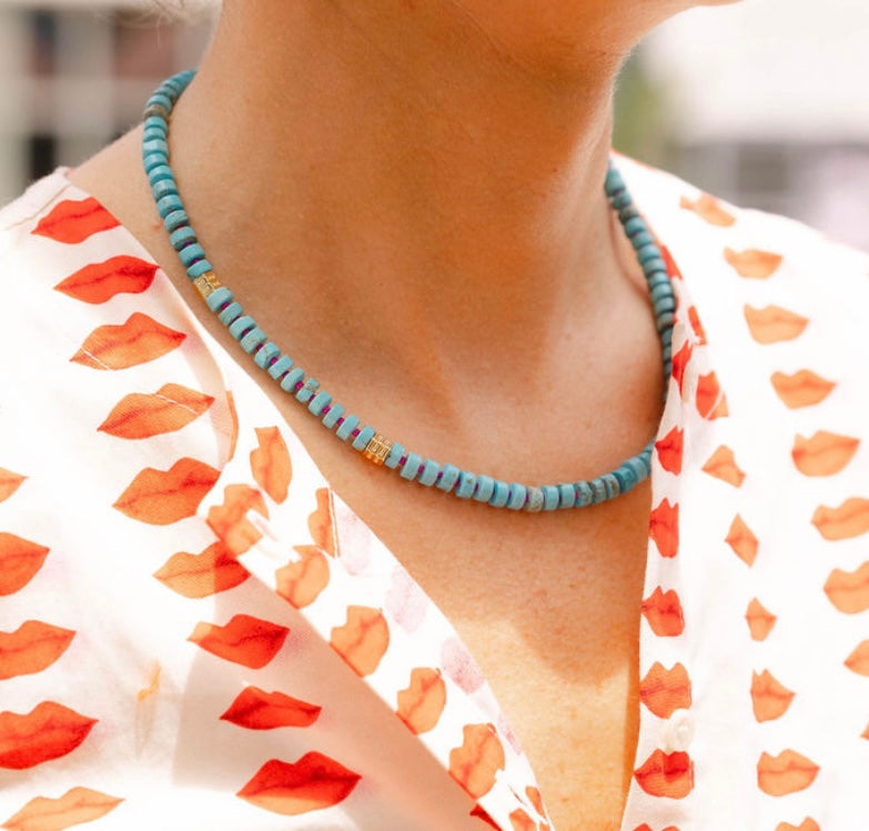 Woman outside wearing a white dress with a red lips print and a blue/gold beaded necklace around her neck