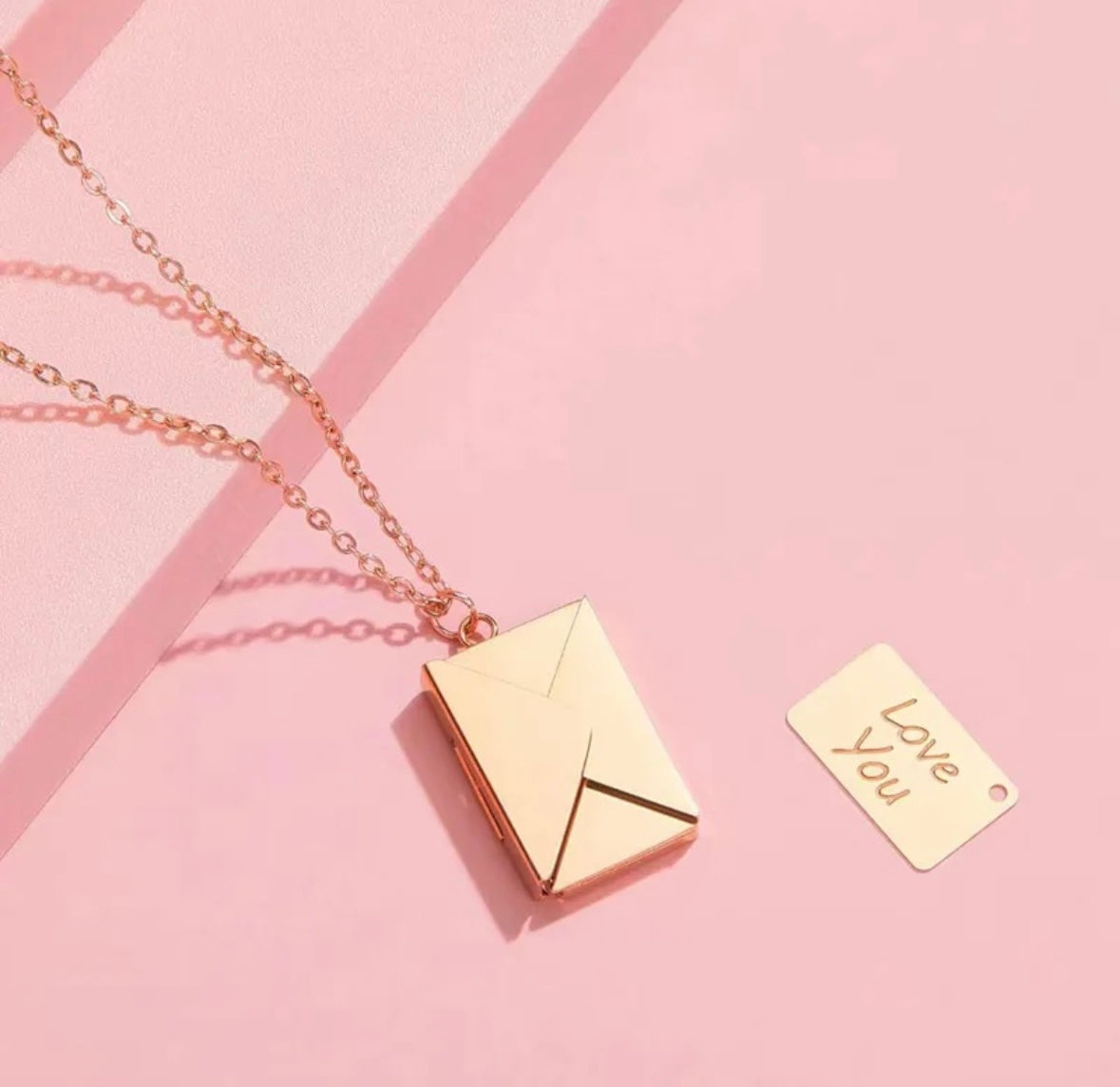 Rose gold envelope-shaped pendant necklace on a pink background with a small gold insert that reads "Love You".