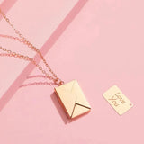 Rose gold envelope-shaped pendant necklace on a pink background with a small gold insert that reads "Love You".