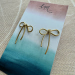 A pair of gold bow earrings on a blue and green card, with the brand mark 'Em' at the top.