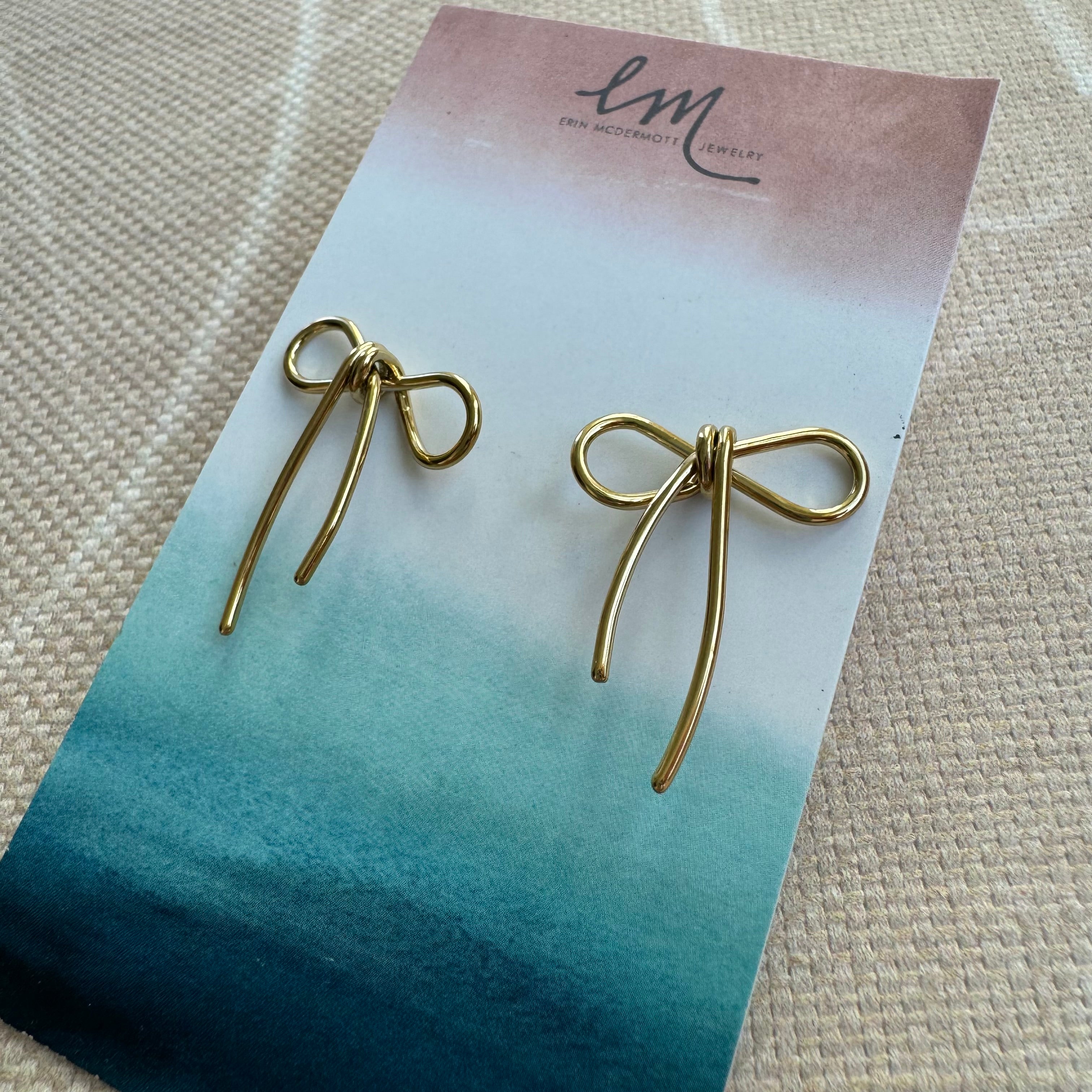 A pair of gold bow earrings on a blue and green card, with the brand mark 'Em' at the top.