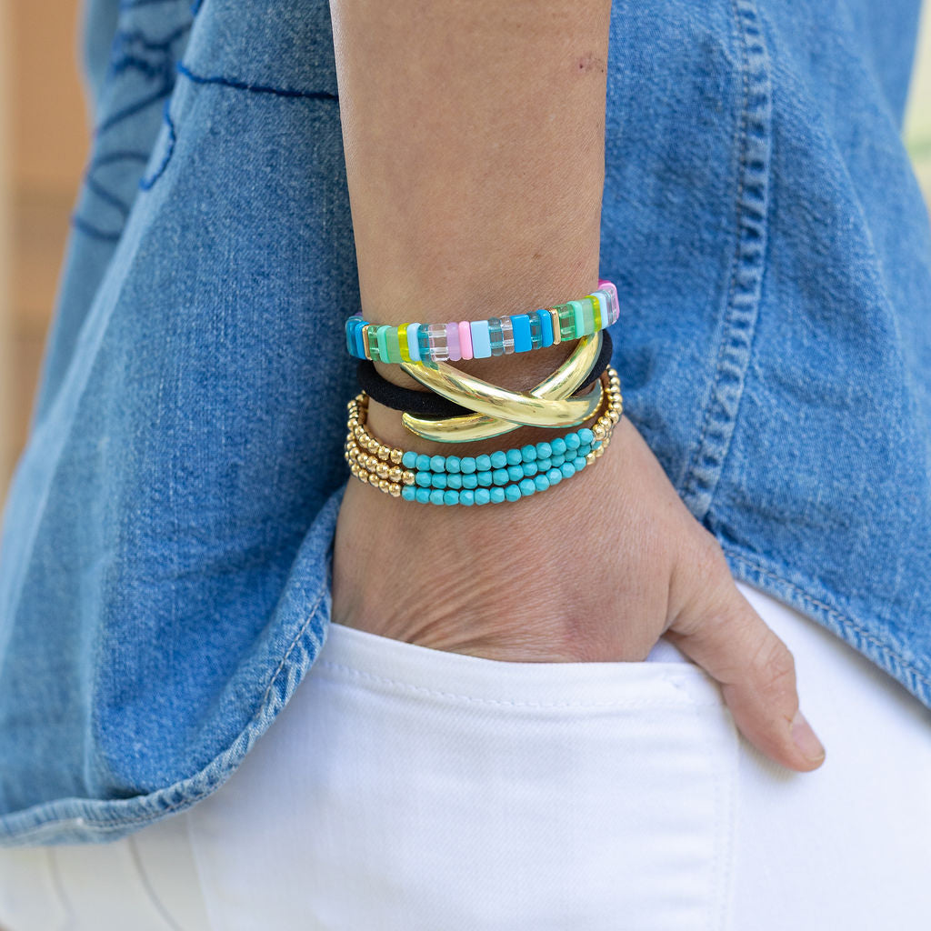 Close-up of a wrist wearing colorful beaded bracelets in pinks, clears, blues and greens. Wrist is also displaying a black wrist band with gold detail and a set of three gold bead bracelets with turquoise bracelets. 