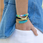 Close-up of a wrist wearing colorful beaded bracelets in pinks, clears, blues and greens. Wrist is also displaying a black wrist band with gold detail and a set of three gold bead bracelets with turquoise bracelets. 