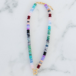 A beaded necklace with a gold toggle clasp, featuring a pattern of multicolored blue and purple hued beads on a marble surface.