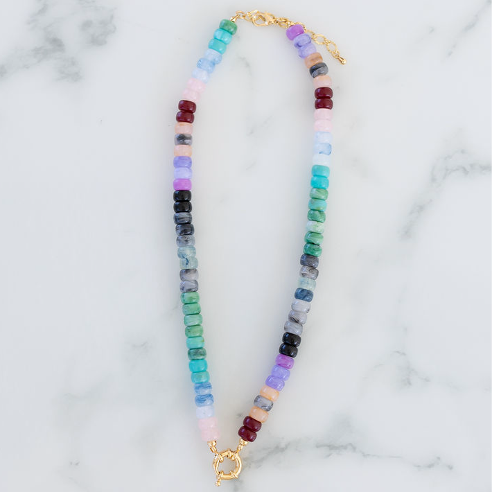 A beaded necklace with a gold toggle clasp, featuring a pattern of multicolored blue and purple hued beads on a marble surface.