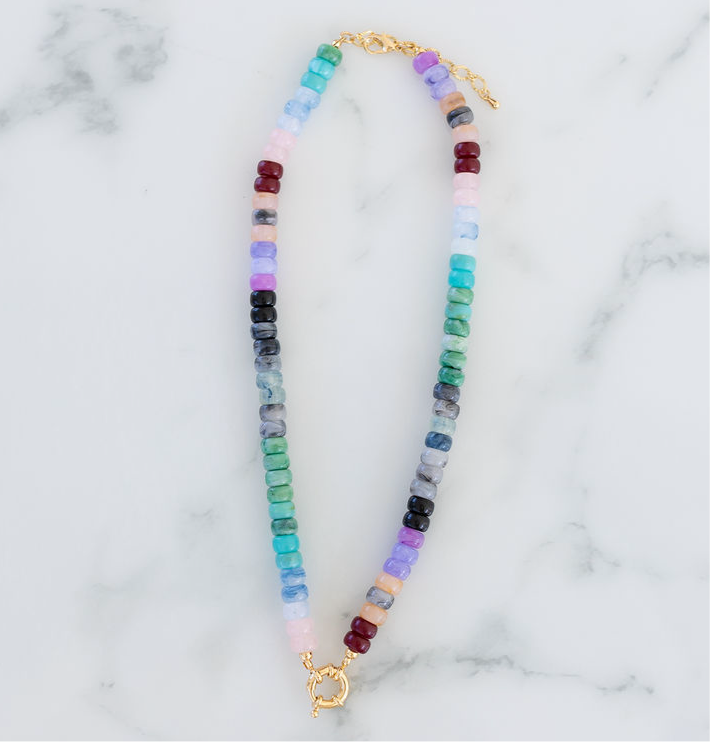 A beaded necklace with a gold toggle clasp, featuring a pattern of multicolored blue and purple hued beads on a marble surface.