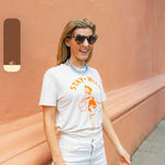Woman wearing sunglasses and a cream shirt with an orange print of a cowgirl with a lasso and the text "Stay Wild" with multiple blue beaded necklaces