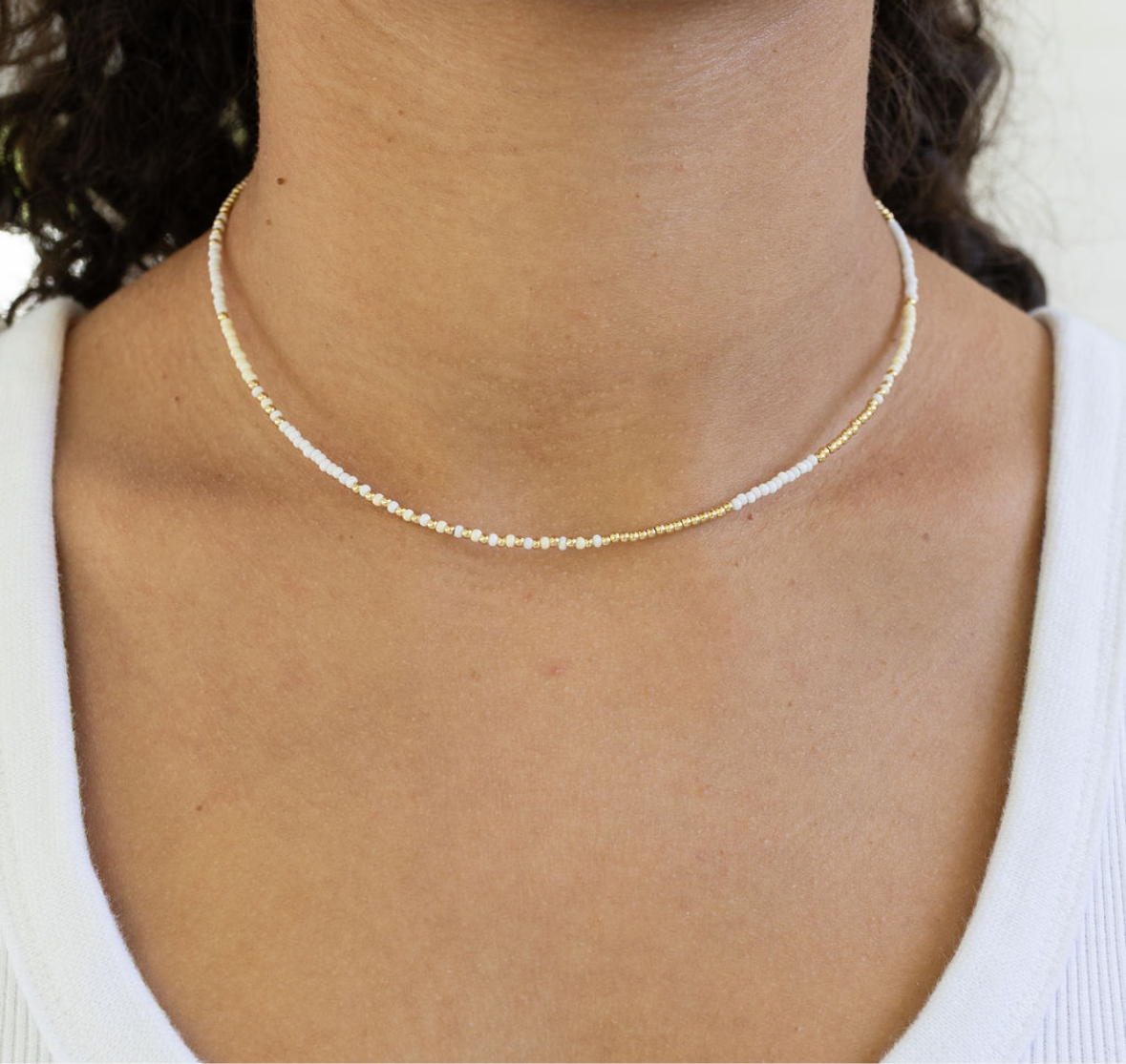 Woman wearing a delicate white seed bead necklace that has gold accent beads.