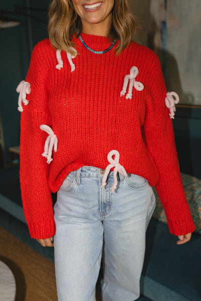 Woman wearing a red knitted sweater with white yarn bows attached and a blue/green beaded necklace