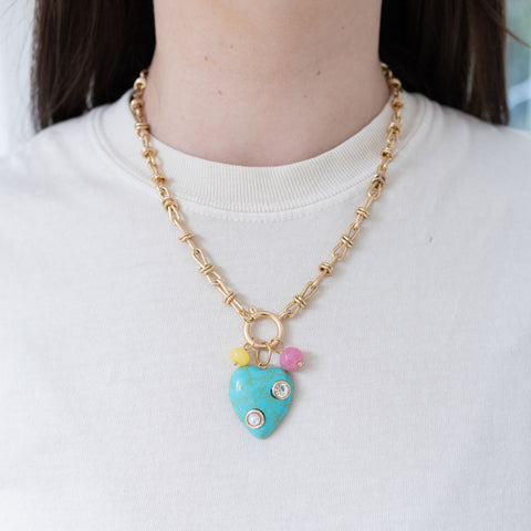 Model wearing a gold toggle chain with a large turquoise heart pendant with diamond and pearl stone inlays. Two small beads, yellow and pink, are separately attached.