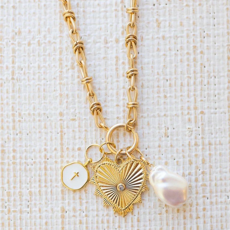 Picture of gold toggle chain with large gold heart pendant with a diamond stone at the center. Also attached is a white disc with gold cross and a fresh water pearl.
