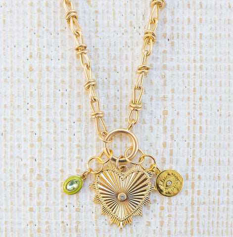 Picture of gold toggle chain with a green stone with green border charm, a gold evil eye charm and a large gold heart pendant with a diamond in the center.