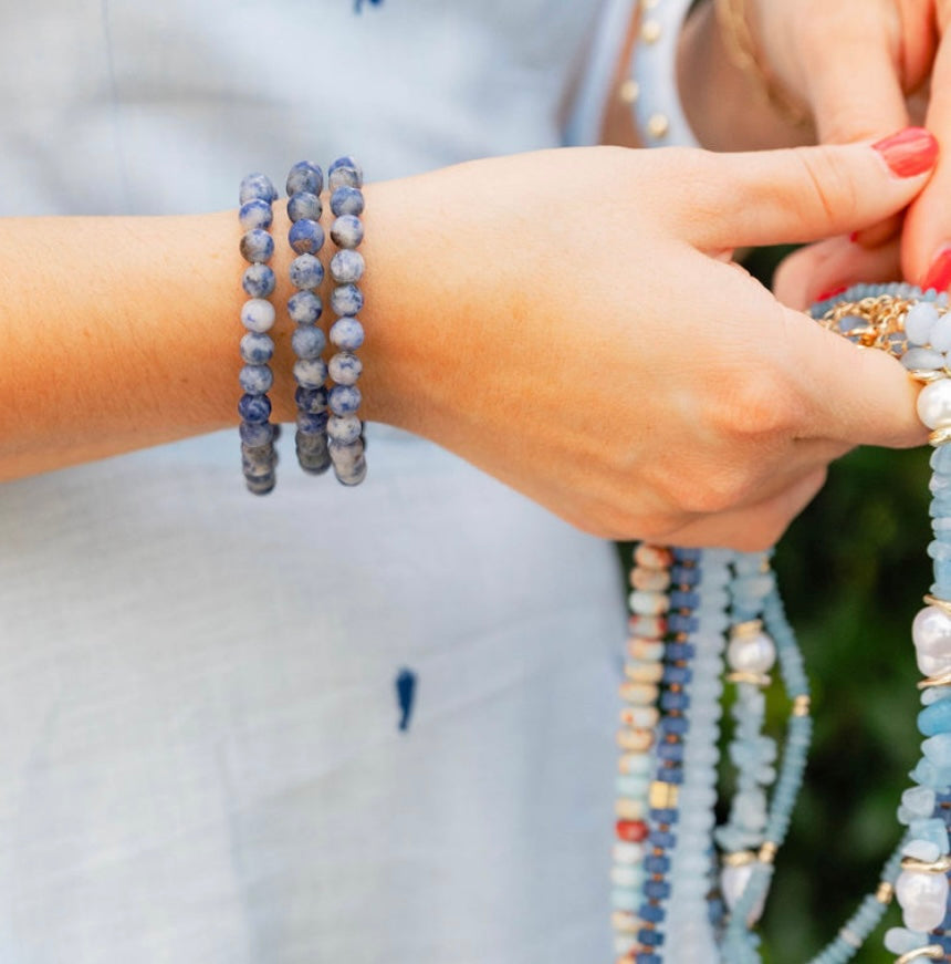Picture of model's hands holding strings of beautiful blue beads. On her wrist are dark blue and gray marbled beaded bracelets.