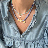 Model in a denim ruffle shirt displaying two necklaces, one a dainty blue bead necklace with a small white disc pendant. The other necklace is made of blue and white striped beads with a tan polka dot fish bead at its center and a small gold disc pendant.