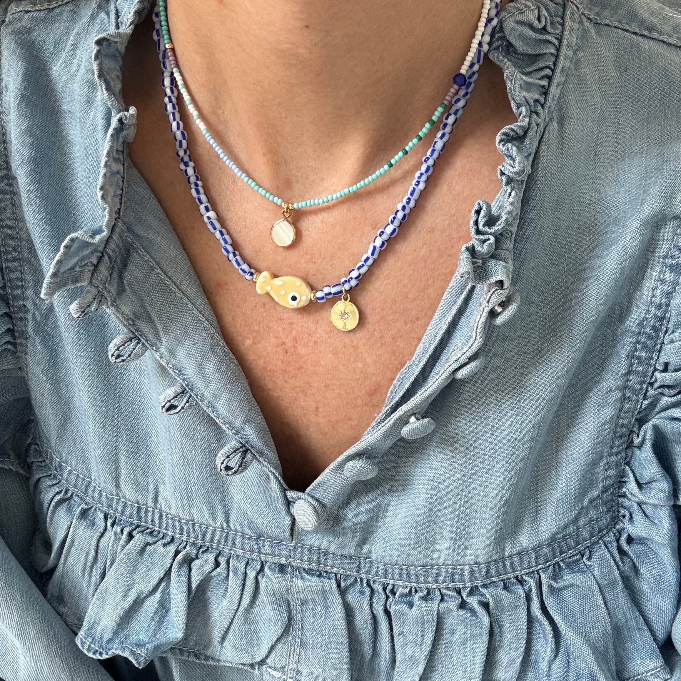 Model in a denim ruffle shirt displaying two necklaces, one a dainty blue bead necklace with a small white disc pendant. The other necklace is made of blue and white striped beads with a tan polka dot fish bead at its center and a small gold disc pendant.