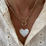 Model wearing a gold toggle chain with a large white heart pendant trimmed with gold studs.