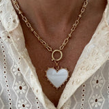 Model wearing a gold toggle chain with a large white heart pendant trimmed with gold studs.