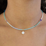 Necklace with tiny blue beads and a small white disc pendant worn by a person, close-up view.