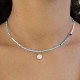 Necklace with tiny blue beads and a small white disc pendant worn by a person, close-up view.