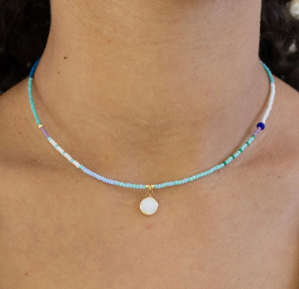 Necklace with tiny blue beads and a small white disc pendant worn by a person, close-up view.