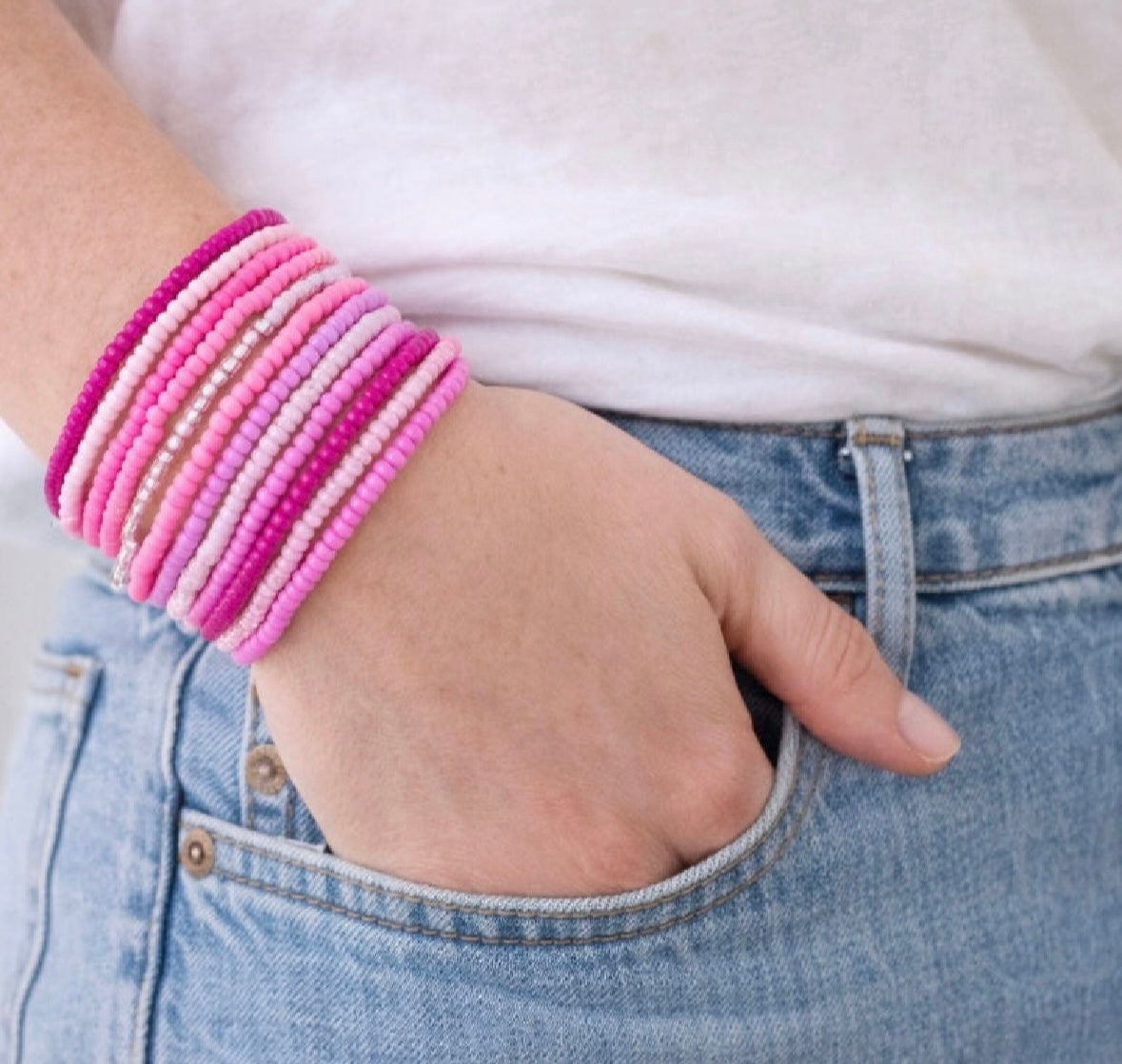 Hand wearing multiple pink beaded bracelets with a white shirt and blue jeans.