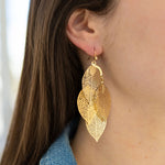 A person wearing gold drop dangle earrings with a geometric leaf design.
