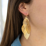 A person wearing gold drop dangle earrings with a geometric leaf design.
