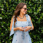 Woman outside in front of a bush in a blue sheer dress wearing sunglasses looking away smiling holding various beaded necklaces in her hands and a blue beaded necklace around her neck