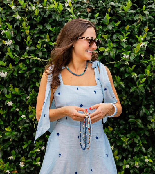 Woman outside in front of a bush in a blue sheer dress wearing sunglasses looking away smiling holding various beaded necklaces in her hands and a blue beaded necklace around her neck